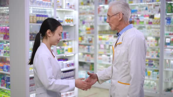 Side View Senior Caucasian Man and Young Asian Woman in Uniform Shaking Hands Talking in Pharmacy