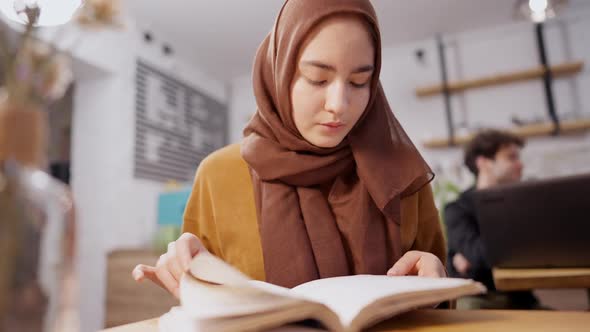 Absorbed Slim Middle Eastern Young Woman Turning Pages of Book Sitting in Cafe