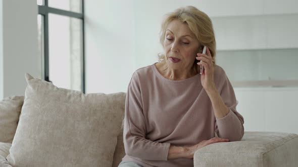 A Sad Elderly Woman is Talking on a Mobile Phone at Home Sitting on the Couch