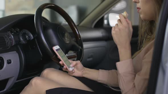 Female Office Worker Sitting in Car, Eating Sandwich and Scrolling on Smartphone