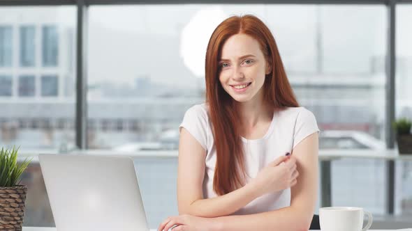 Portrait of Happy Redhead Girl Sitting at Table with Laptop Looking at Camera and Smiling