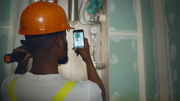 Back View of African Engineer with Flashlight Taking Photo on Smartphone of Broken Heating System