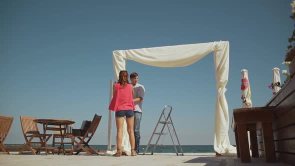 Caucasian Female and Male Decorators Installing and Decorating Wedding Arch on Terrace Near Sea in