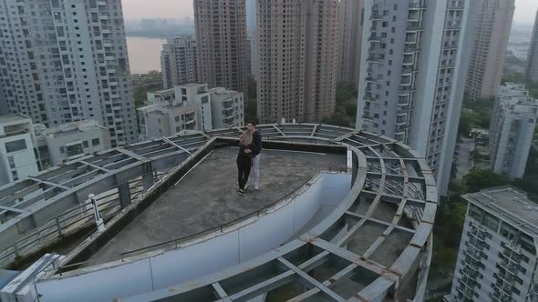 Aerial Orbital Shot of Hugging Couple on the Roof Top of Modern Apartments Building on the Sunset