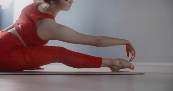 Young Woman in Red Tracksuit Sitting on the Yoga Mat and Leaning Forward To Her Feet, Person Does