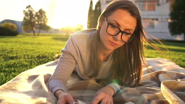 Girl in Glasses Reading Book Lying Down on a Blanket in the Park at Sunset