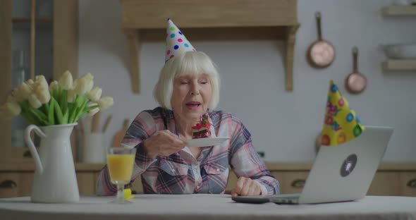 Senior Woman Celebrates Birthday Online with Piece of Cake and Candle. 70s Woman in Birthday Hat