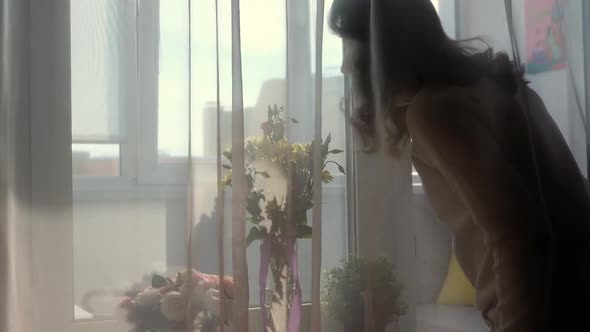 Young Woman Smelling Spring Flowers in Vase and Looking at Window From Home