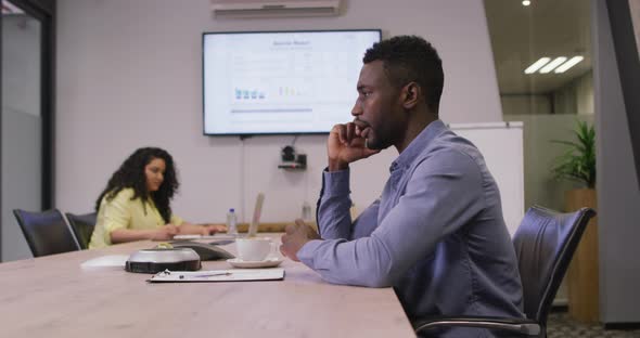 African american businessman sitting at desk, talking on smartphone in modern office