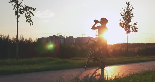 Woman cyclist is drinking water after bike ride in park against sunset. Triathlon concept