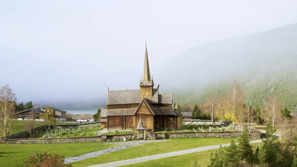 Wooden Exterior Facade Of Lom Stave Church Against Misty Forest Mountains In Lom, Norway. Aerial Til