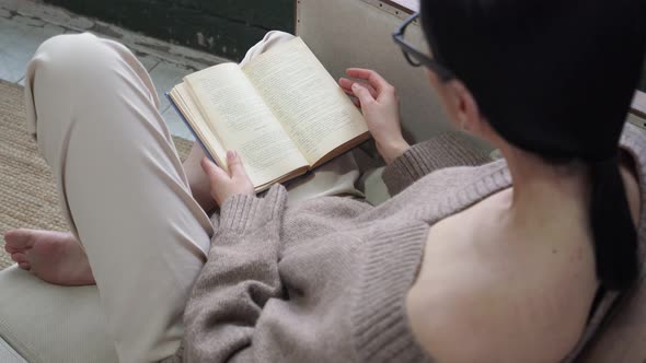 a Young Woman in Cozy Clothes and a Smooth Hairdo is Reading a Book While Sitting on the Couch with