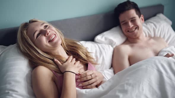 Young Couple Enjoying Time at Home Together Lying in Bed in Bedroom Looking at Each Other Laughing