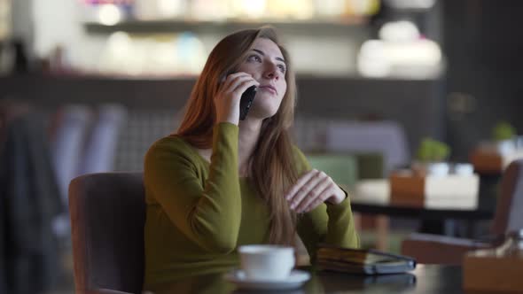 Charming girl is sitting in a cafe indoors and talking on the phone. Girl has a cup of coffee and a