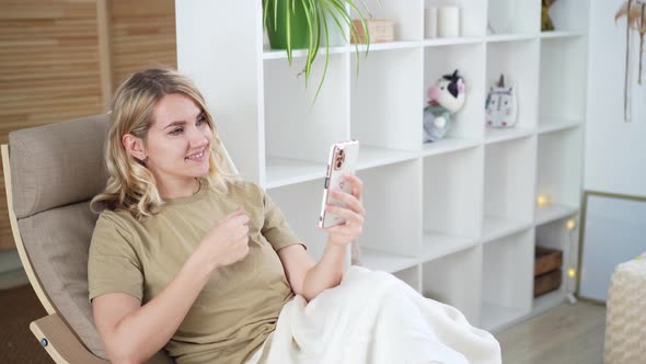 Cute young blonde woman uses phone while sitting at home in armchair