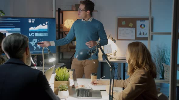 Young Employee Making Presentation to Group of Colleagues in Dark Office Speaking and Pointing at