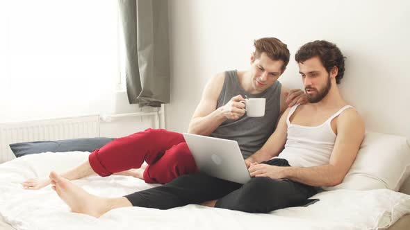 Happy Young Gay Couple of Men Are Using Modern Laptop Computer Lying in Bed in the Bedroom.