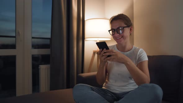 Woman with Glasses Sitting on the Couch in a Cozy Room and Using Smartphone for Surfing