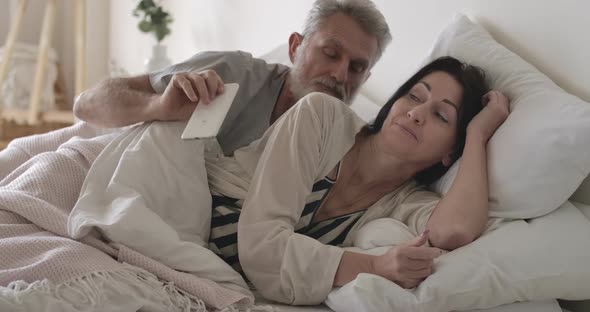 Furious Caucasian Senior Man Taking Away Wife's Smartphone in Bed. Husband Looking Angrily at