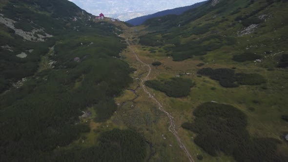 Aerial view of a hiking path in a lush green area, hills left and right and a building in the backgr