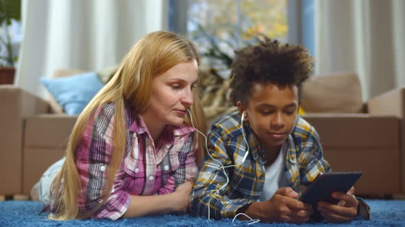 Happy Foster Mother and Son Lying on Floor and Using Tablet Pc and Headphones at Home