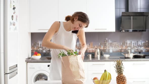 Beautiful Young Caucasian Female Coming to Kitchen with Grocery Shopping Bag Talking on Mobile Phone