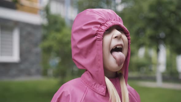 Funny Little Girl in Pink Coat and Hood Catching Rain Drops with Tongue. Portrait of Cheerful