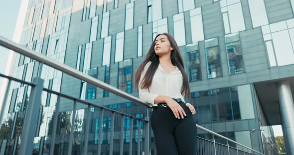 Beautiful Attractive Elegantly Dressed in White Shirt Woman Stands at Railing Next to Glass Building