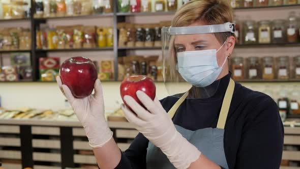 A Saleswoman in a Medical Mask Holds an Apple in Her Hands in a Grocery Store