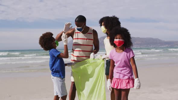 African american parents with two children wearing face masks collecting rubbish from the beach