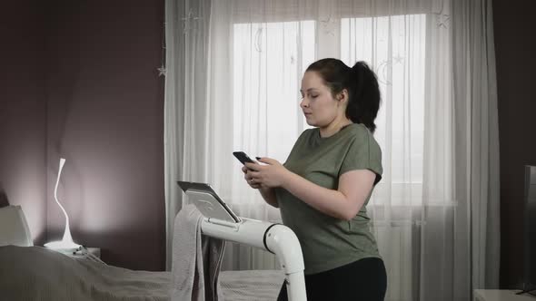 Portrait of woman with smartphone in hands is walking on treadmill at home