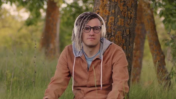 Young Relaxed Man in Glasses Headphones Sitting Near Tree in Green Grass Listening to Music on