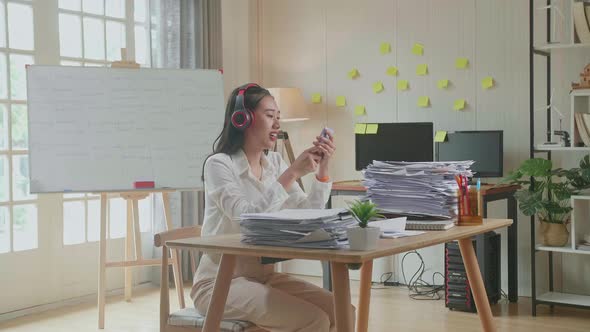 Asian Woman With Headphones Listening To Music On Smartphone After Working With Documents