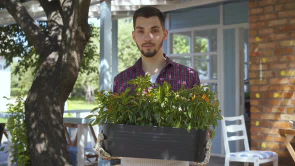 Portrait of Young Bearded Farmer Holding a Pot of Flowers in Hands Looking at Camera Smiling