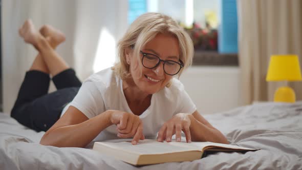 Beautiful Aged Woman Reading Book While Lying on Bed with Grey Linen at Home