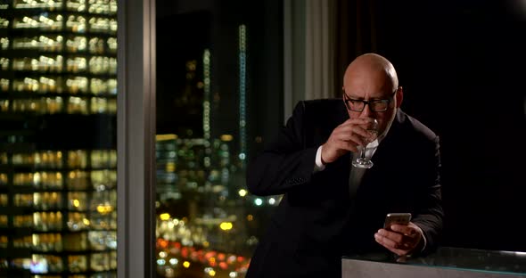 Businessman in Glasses Drinks Wine and Checks His Phone at a Large Window. Outside the Window Is