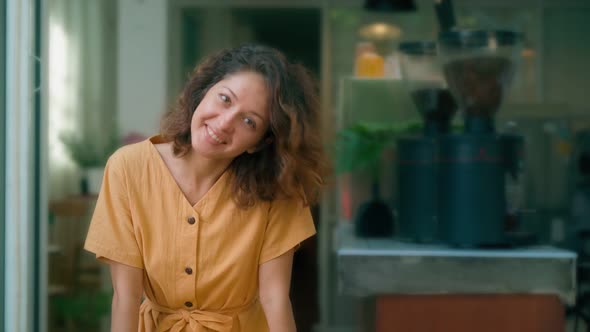 Smiling Curly Woman in Yellow Dress Showing Tongue Dancing Happily While Sits in Coffee Shop