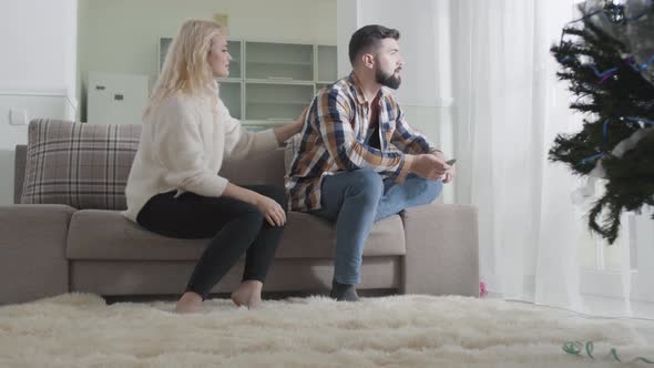 Young Blond Woman Caressing Man's Back and Talking. Offended Bearded Guy Sitting on Sofa