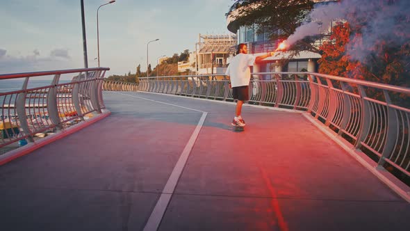 Back View of Young Guy Riding Skateboard with Signal Fire on Bridge