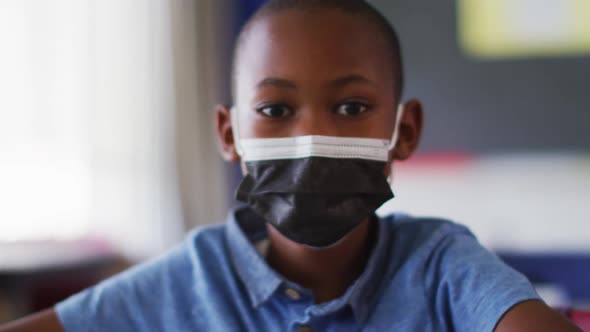 Portrait of african american schoolboy wearing face mask, sitting in classroom looking at camera