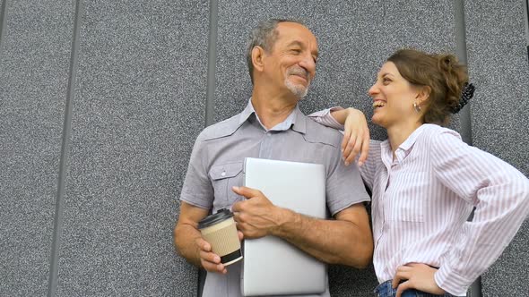 Young Woman and Senior Man are Waiting for a Meeting Standing Near the Grey Wall Outdoors in the