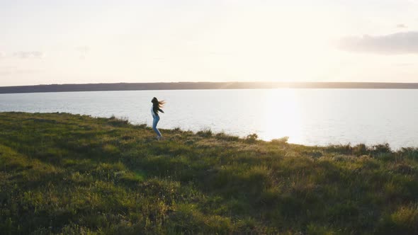 Tracking Aerial Shot of Young Woman Enjoying Sunset and Having Some Fun in Field Near the Cliff