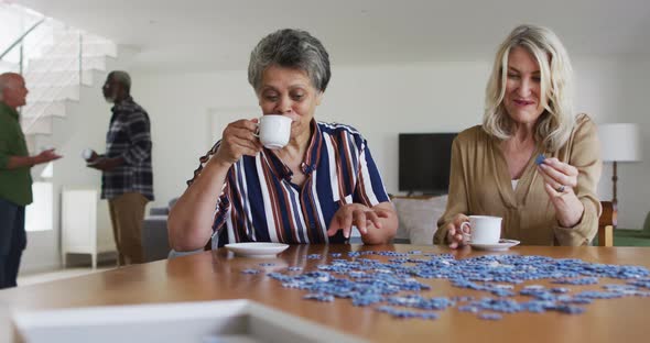 African american and caucasian senior women sitting by table doing puzzles drinking tea