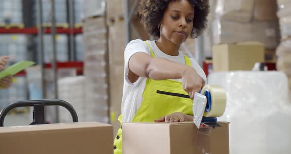 African Female Worker Packing Cardboard Box with Tape Gun Dispenser in Warehouse