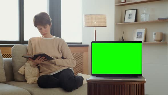 A Laptop with a Green Screen Stands on a Table Against of a Woman Reading a Book