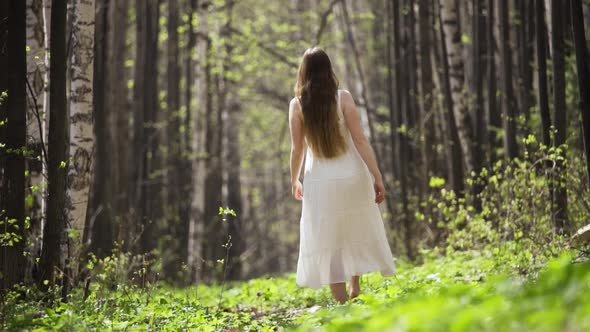 A Beautiful Romantic Woman with Loose Long Hair in a Light White Dress Walks Barefoot Through a