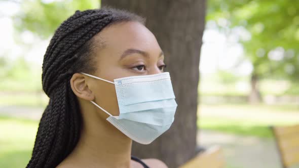 A Young Black Woman in a Face Mask Looks Around and Sits on a Bench in a Park on a Sunny Day