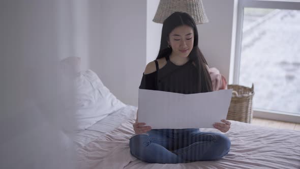 Wide Shot Front View of Positive Smiling Young Woman Analyzing Business Project Sitting on Bed in