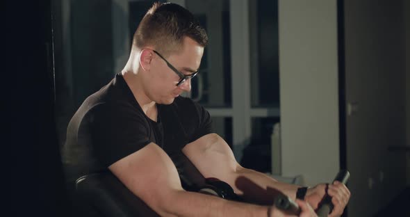 Athlete with Glasses Works Out with the Simulator to Pump Biceps in a Gym