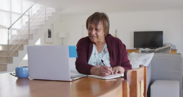 Smiling african american senior woman sitting at dining table, using laptop and writing in notebook